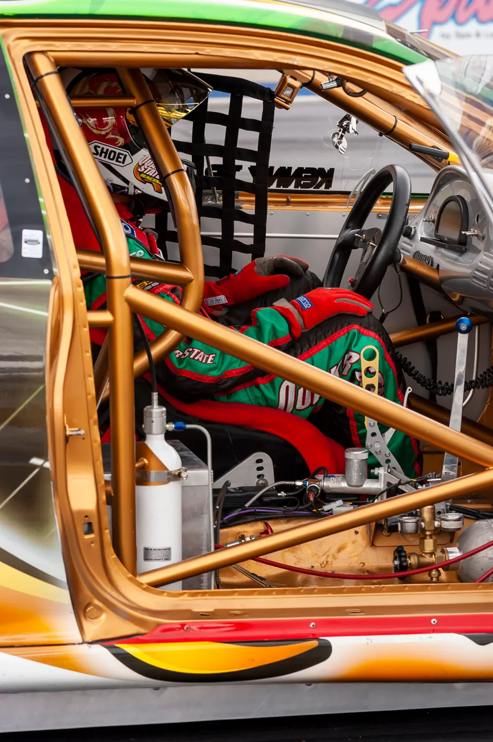 A detailed view of a race car's interior, showing the driver in a racing suit, gold roll cage, and safety equipment.