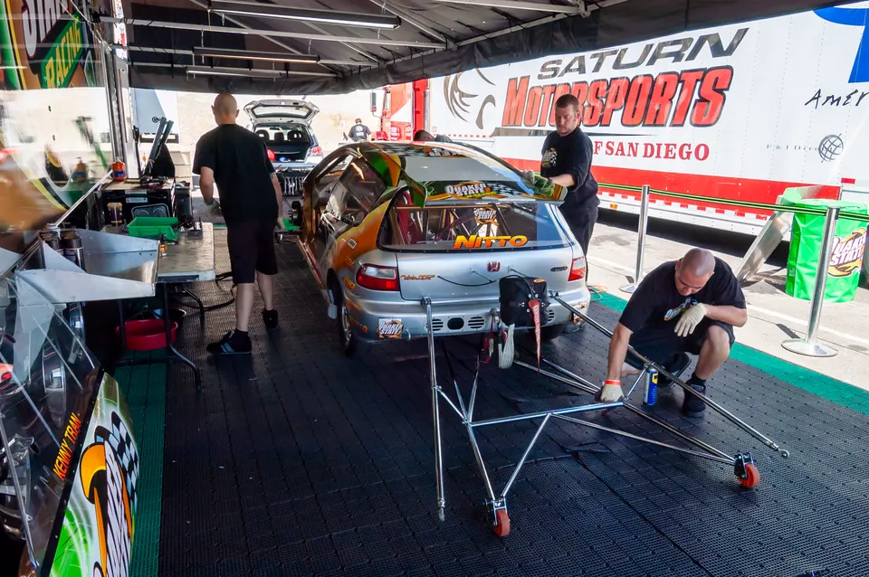 A drag racing Honda Civic in a pit area with mechanics working on its wheelie bars, next to a Saturn Motorsports trailer.