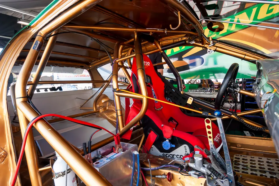 Detailed view of a race car interior featuring a gold roll cage, red Sparco racing seat, and safety equipment.
