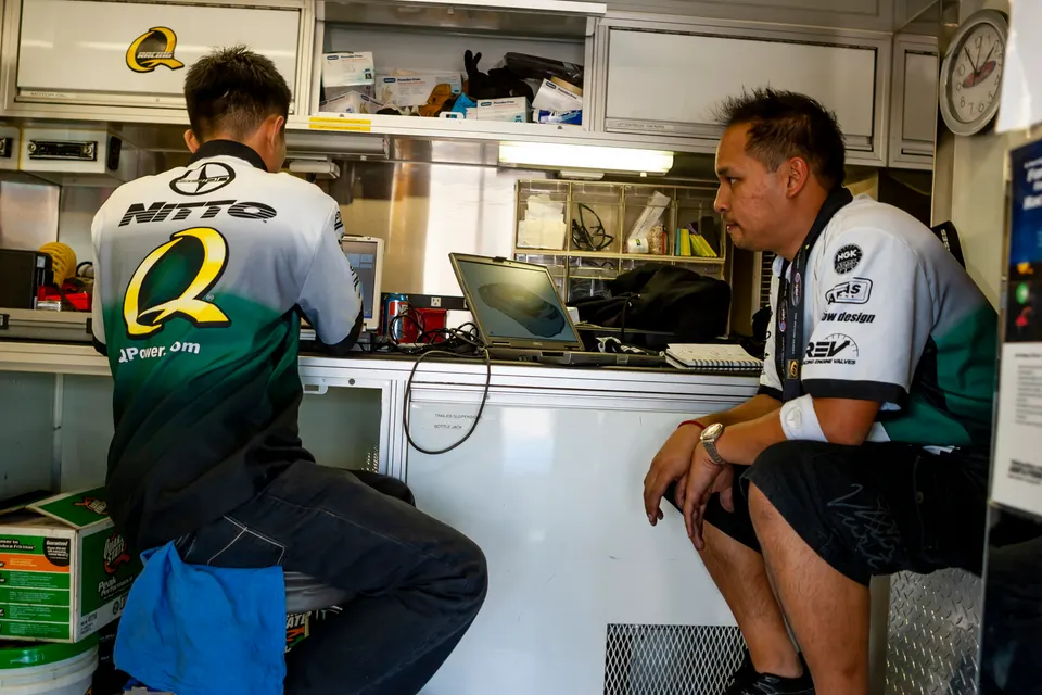 Two mechanics in a shop trailer, one working on a laptop displaying a 3D model of an induction part, the other observing.