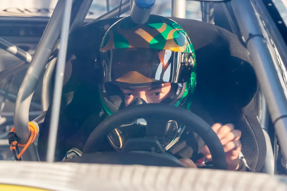 A race car driver in a green and brown camouflage helmet and racing suit, gripping the steering wheel inside a roll-caged cockpit.