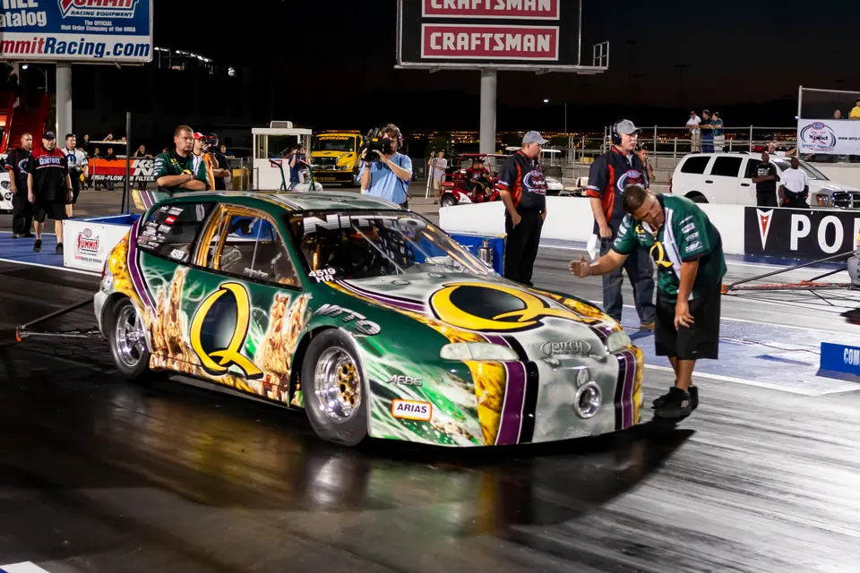 A custom-painted Honda Civic drag car in the staging lane at a night race event with crew members and spectators.