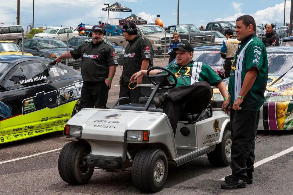 Pit crew members and driver Shawn Hillier at a drag racing event with a custom race car and golf cart.