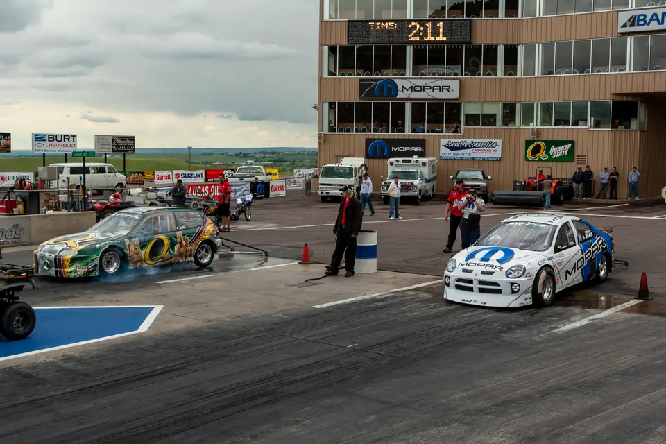 Two drag racing cars, a white Dodge Neon SRT-4 and a green hatchback, at the starting line of a drag strip.