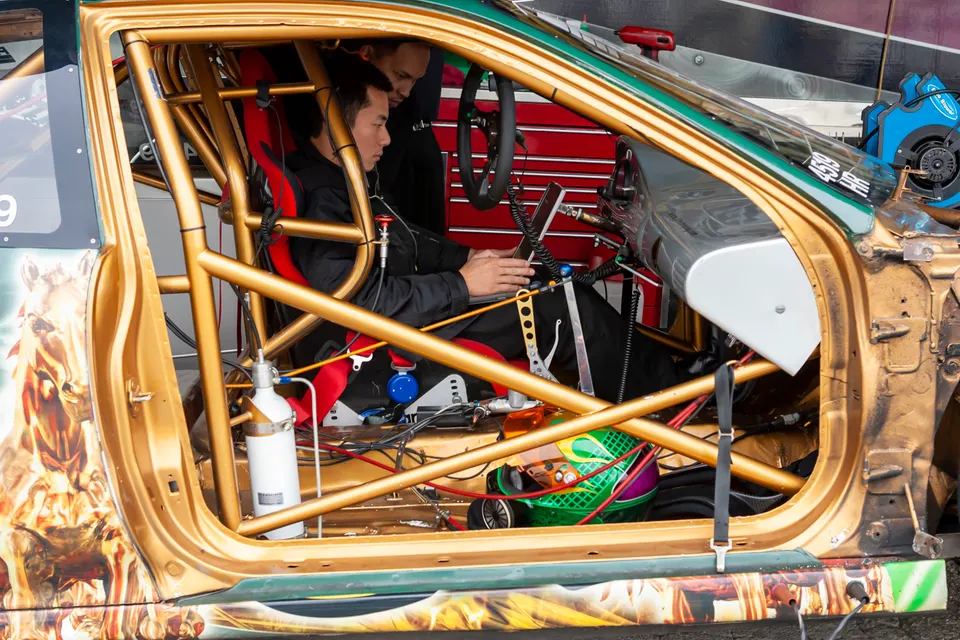 Driver in a gold race car with a prominent roll cage, using a laptop for tuning while a crew member looks on.
