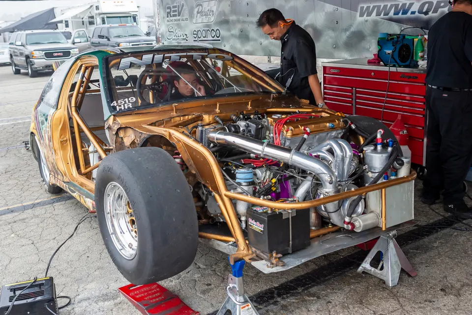 A gold Honda Civic drag car with its front end removed, showing the turbo engine, being prepped by mechanics at a race track.