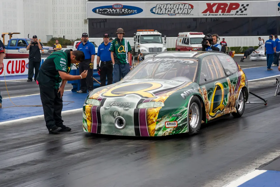 A highly modified green and silver Honda Civic drag race car on the starting line at Pomona Raceway with crew members.