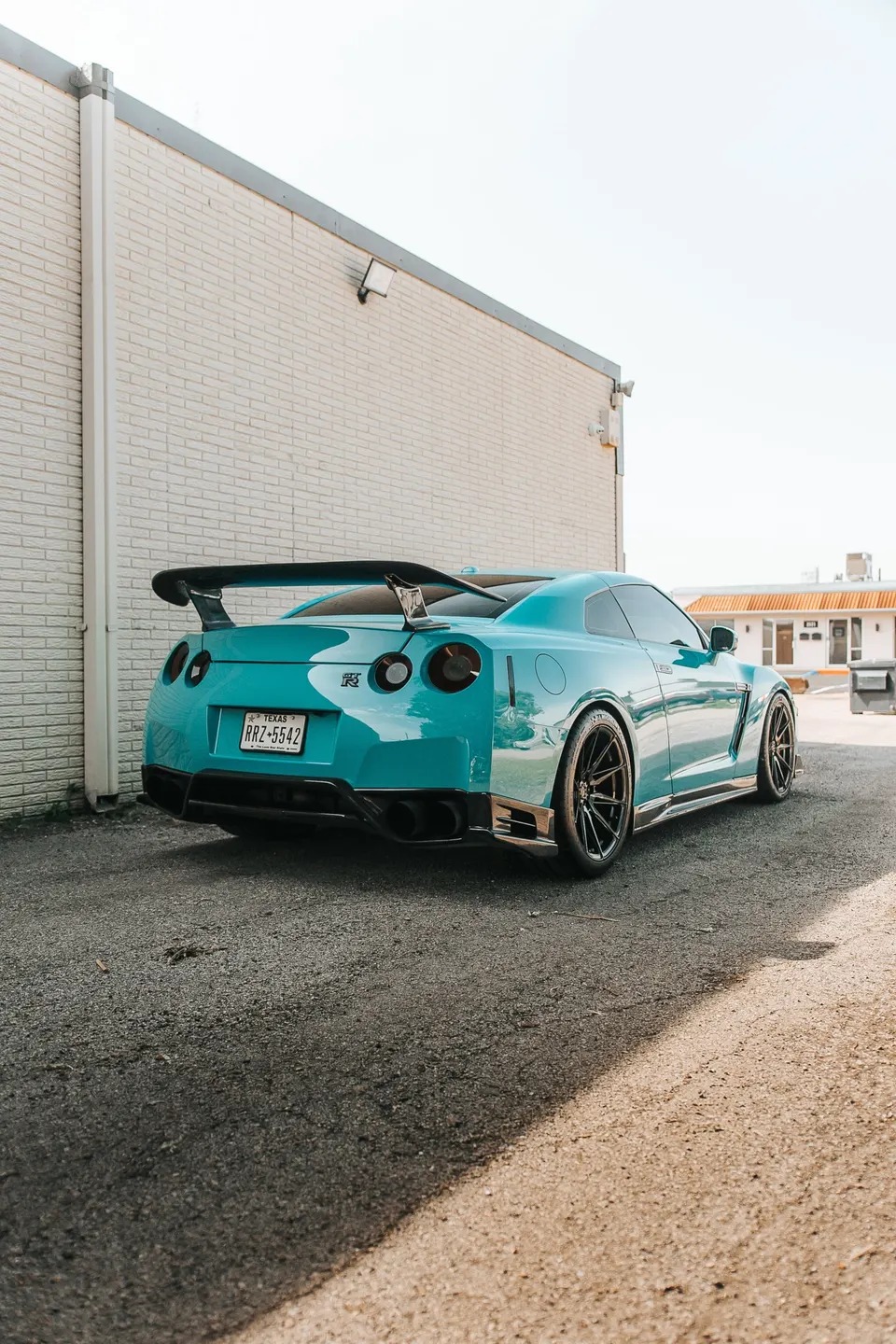 Turquoise Nissan GT-R R35 with a large carbon fiber wing and aftermarket wheels parked next to a brick building.