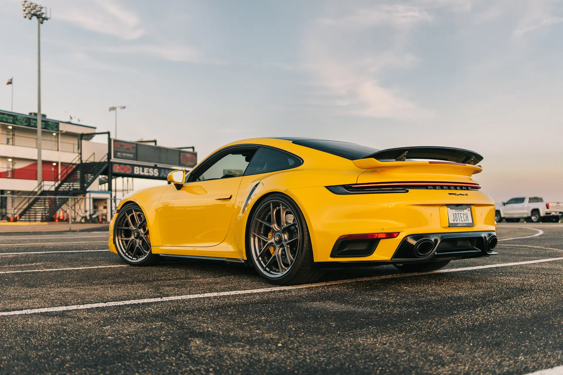 A yellow Porsche 911 Turbo S with aftermarket wheels parked at a drag strip with a building in the background.