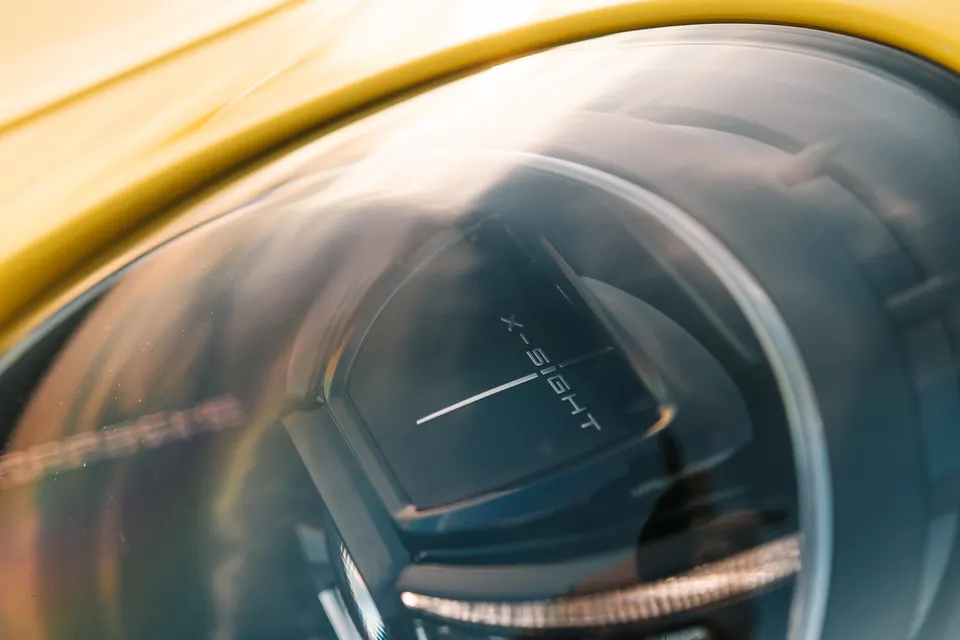 A close-up detail shot of a yellow car's headlight, featuring 'X-SIGHT' branding within the lens.