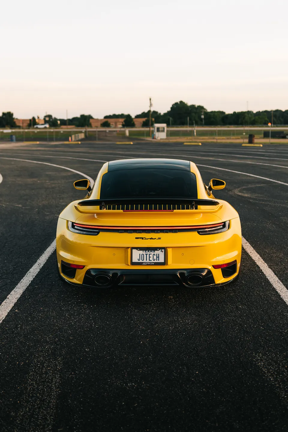 Rear view of a yellow Porsche 911 Turbo S with a Jotech license plate, parked on an asphalt lot.