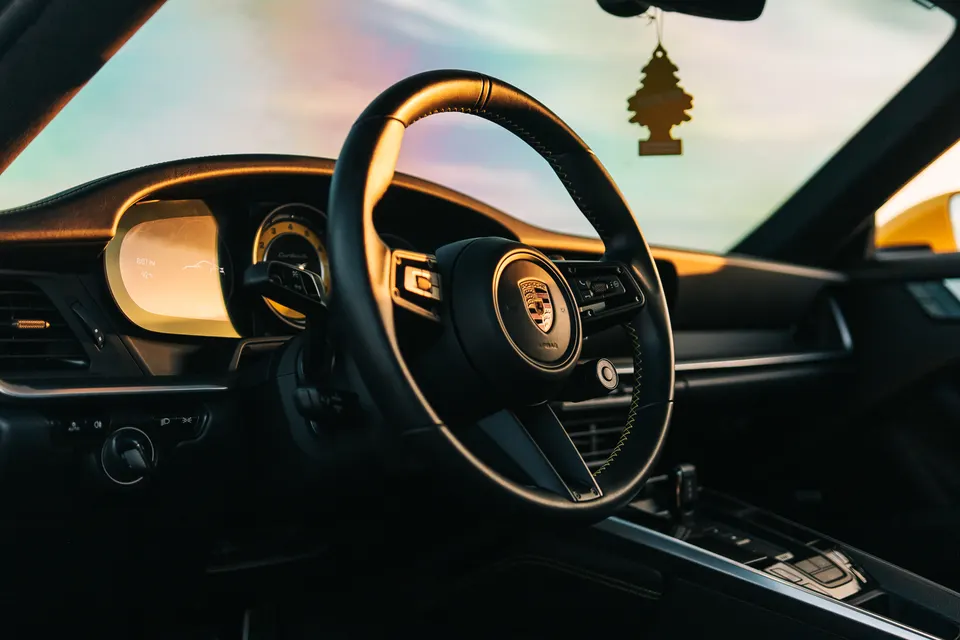 Interior view of a Porsche with a leather steering wheel, dashboard, and an air freshener hanging from the rearview mirror, bathed in golden light.