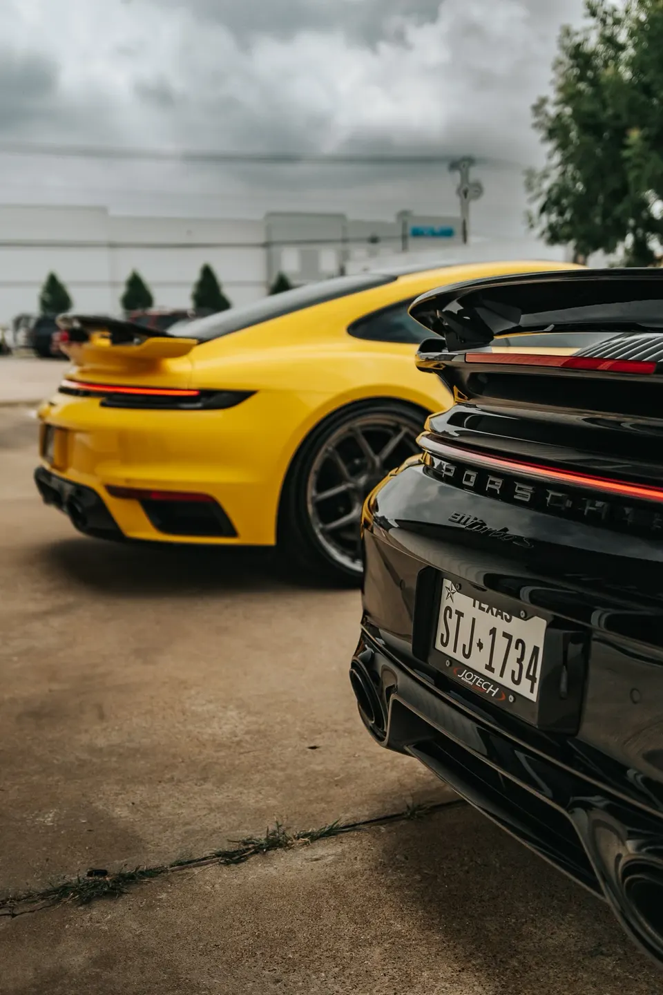 Rear view of a black Porsche 992 Turbo S and a yellow Porsche 992, showcasing their aggressive rear styling and exhaust.