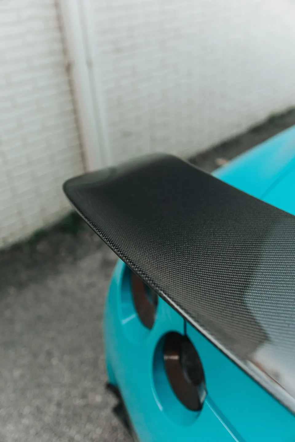 A close-up of a carbon fiber rear wing on a turquoise sports car.