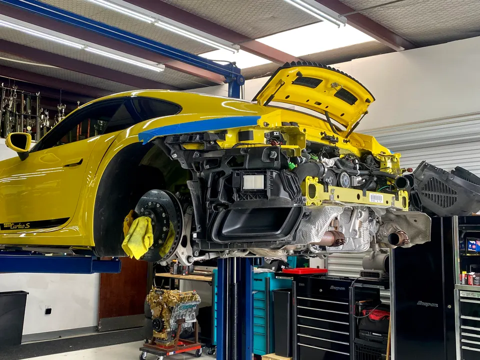 A yellow Porsche 911 Turbo S on a lift in a shop with its rear bumper removed, exposing the engine and exhaust system.