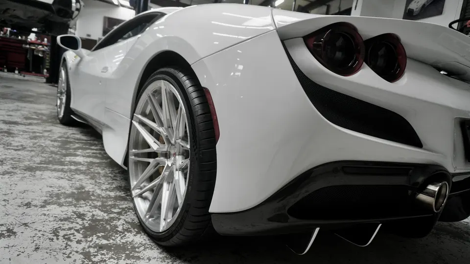 White Ferrari F8 Tributo with aftermarket multi-spoke wheels and carbon fiber rear diffuser in a performance shop.