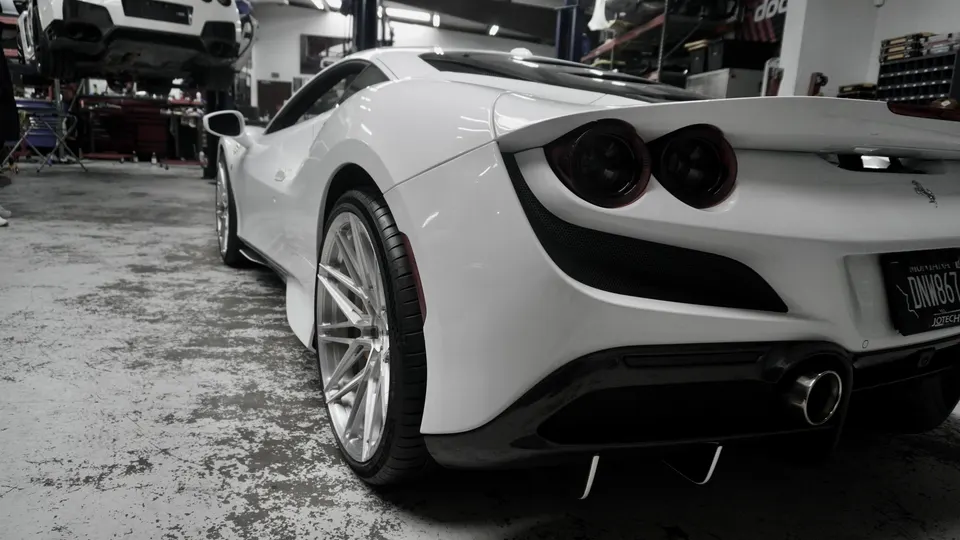 Rear view of a white Ferrari F8 Tributo with custom wheels and aftermarket exhaust in a performance tuning shop.
