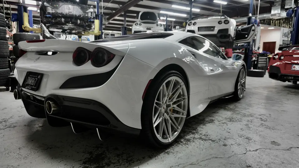 A white Ferrari F8 Tributo with aftermarket wheels and exhaust tips parked in a performance tuning shop.