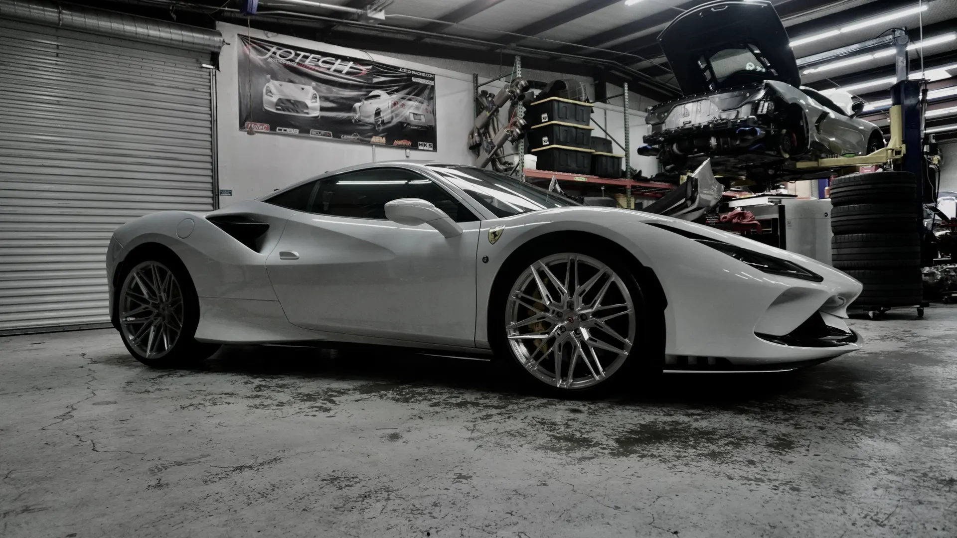 A white Ferrari 488 parked in a performance tuning shop with another car on a lift in the background.