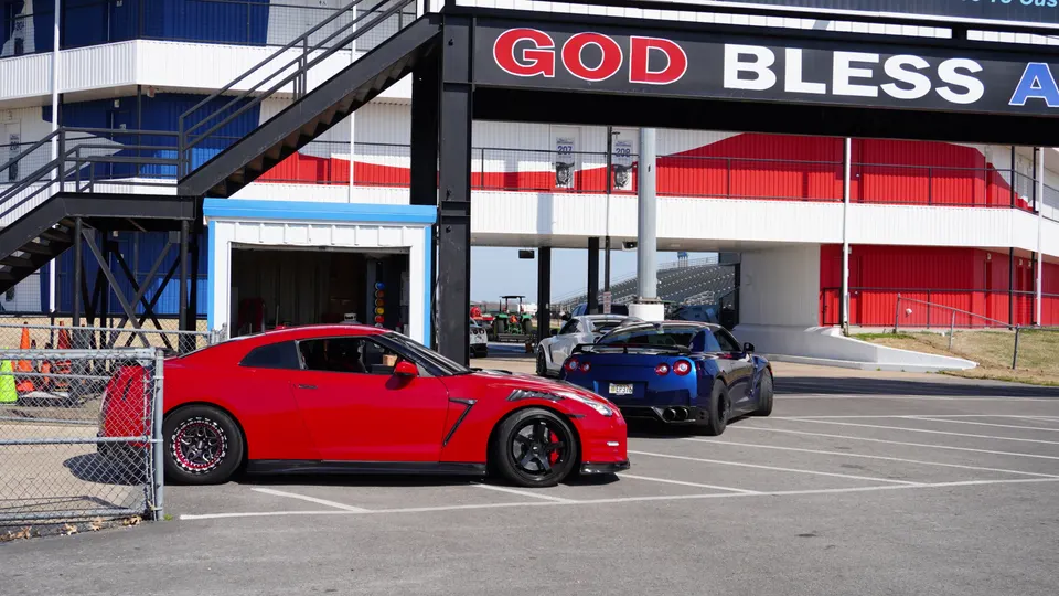 Red and blue Nissan GT-Rs parked at a drag strip facility with grandstands and a building in the background.