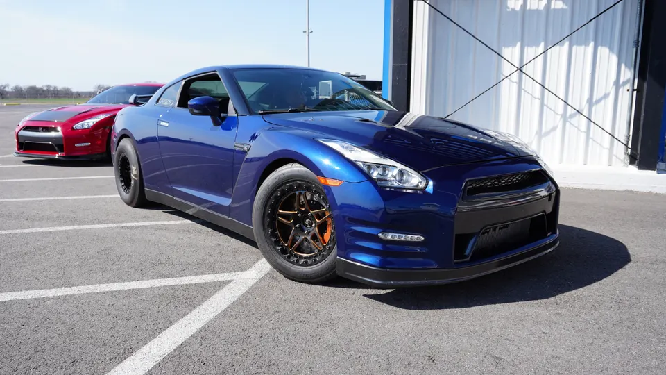 A blue Nissan GT-R with aftermarket wheels parked in a lot, with a red Nissan GT-R in the background.