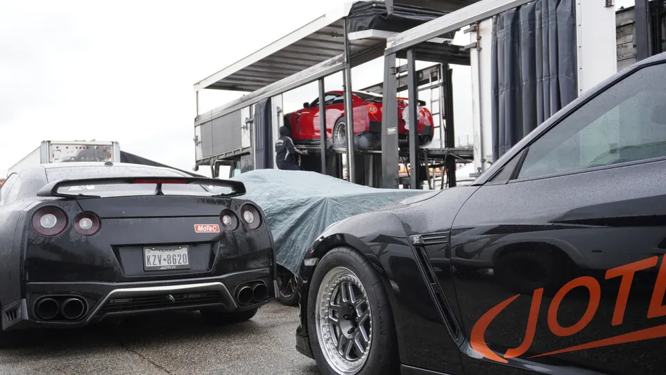 Multiple Nissan GT-Rs in a paddock area, with one black GT-R in the foreground and a red GT-R on a lift.