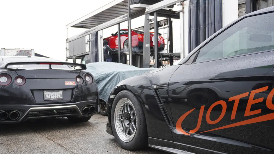 Multiple Nissan GT-R performance cars, including a black Jotech GT-R with aftermarket wheels, at a track event with trailers.