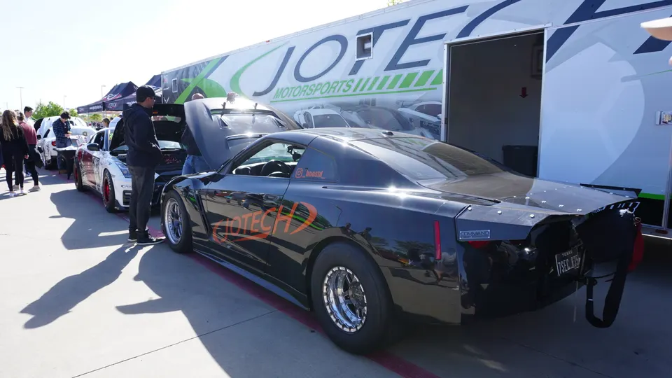 A black drag-spec Nissan GT-R and other modified cars parked next to a Jotech Motorsports trailer at a car event.