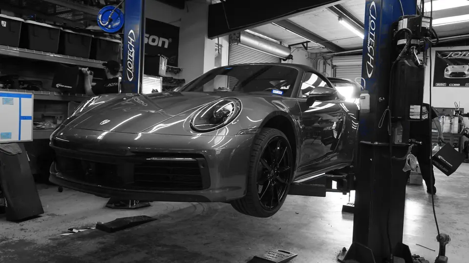 A grey Porsche 911 Carrera on a lift in the Jotech Motorsports workshop, with a technician in the background.