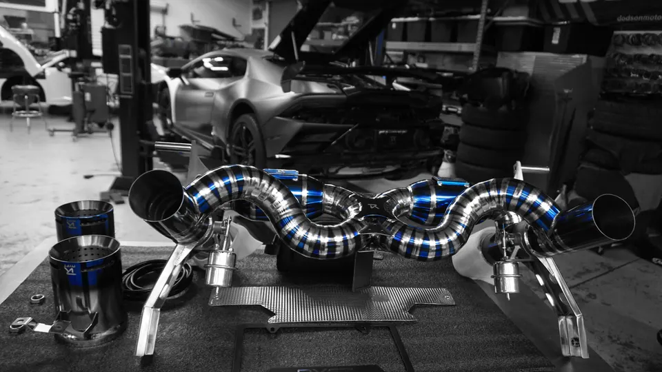 A performance exhaust system with blue accents on a table in a shop, with a Lamborghini Huracan on a lift in the background.