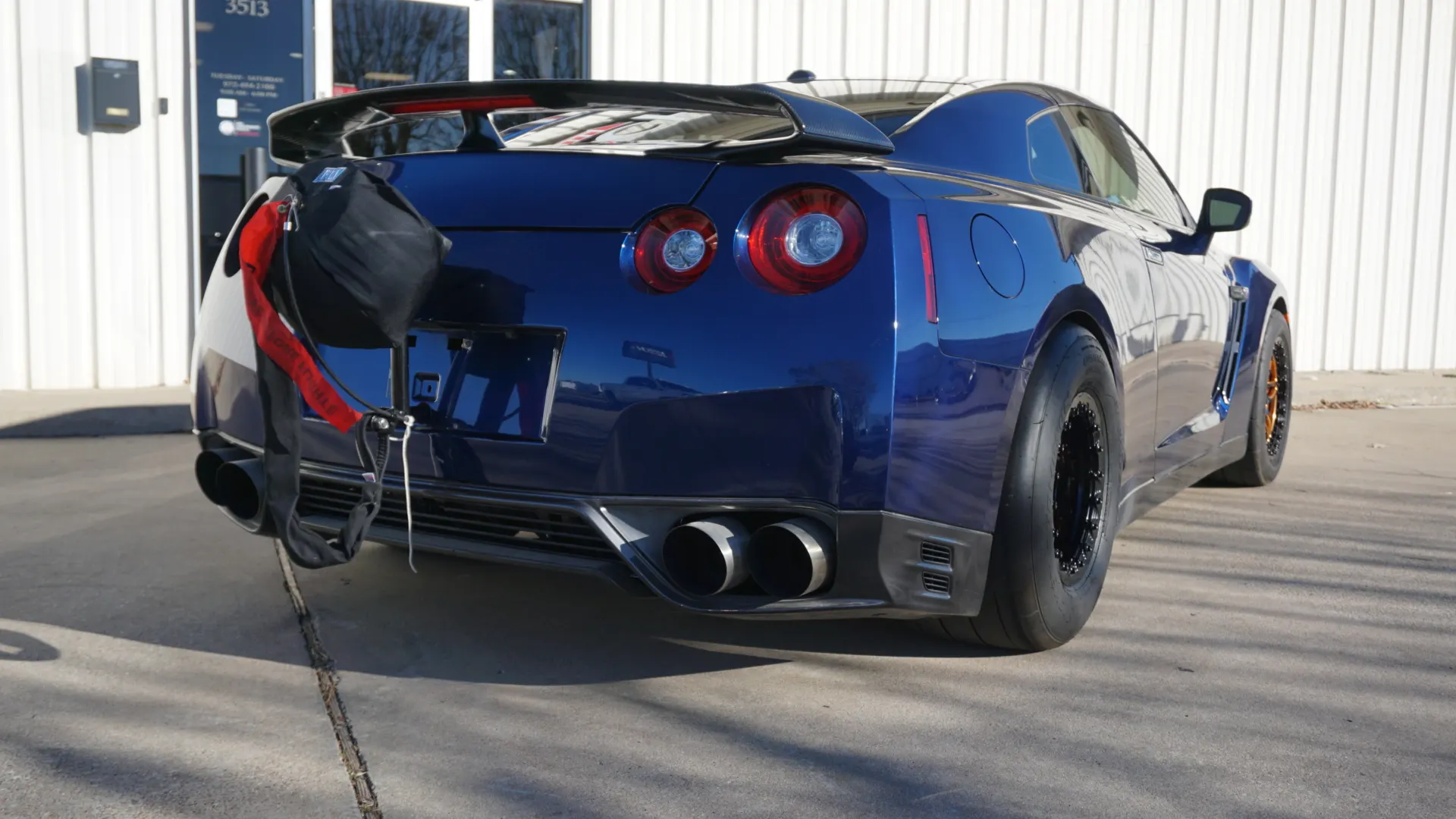 Rear view of a blue Nissan GT-R R35 drag car with a parachute, aftermarket exhaust, and drag slicks parked outside a building.