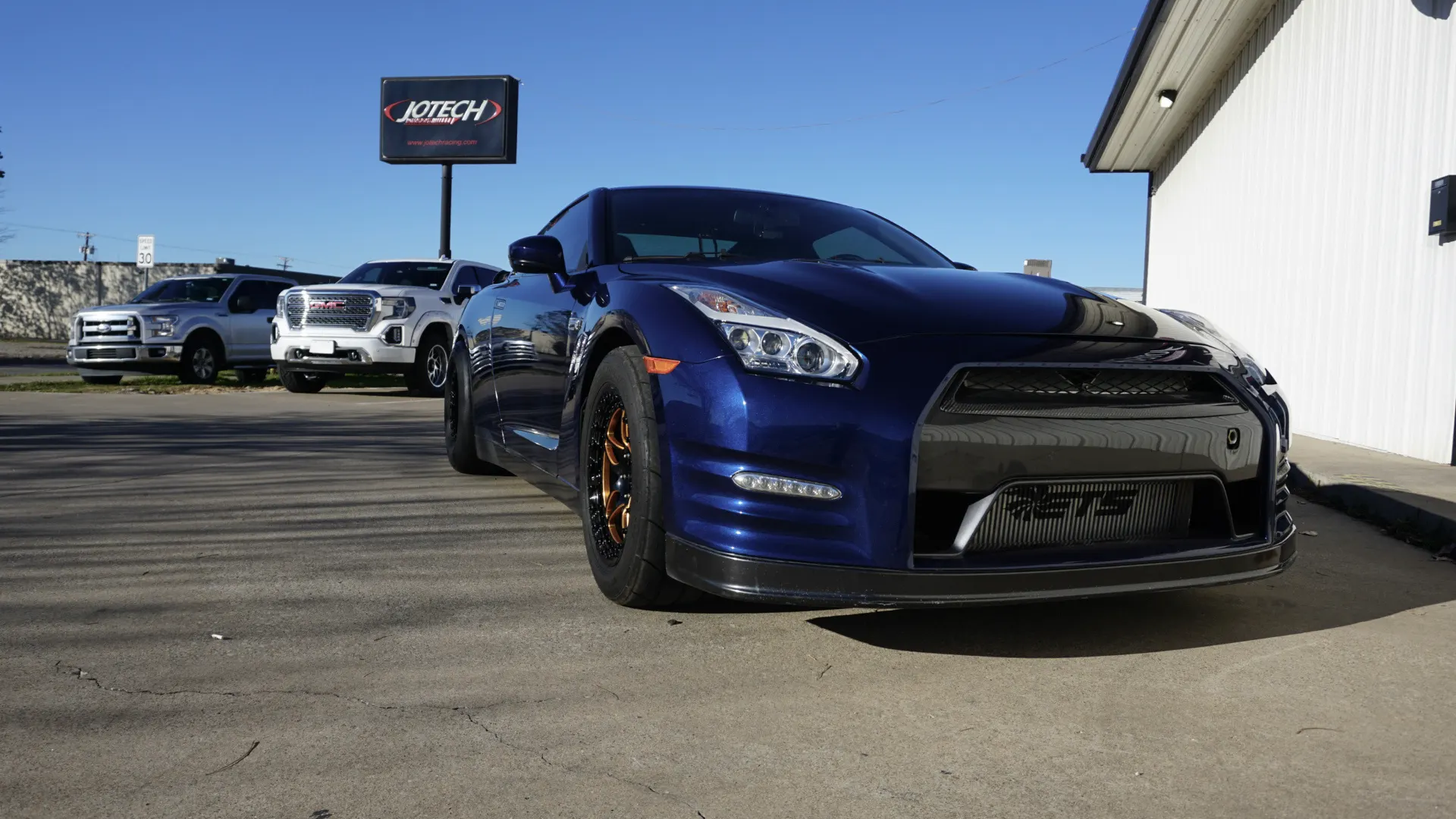 A blue Nissan GT-R with aftermarket wheels and a front splitter parked outside Jotech Motorsports.