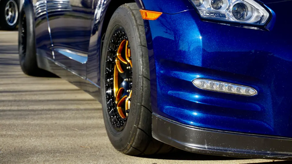 A close-up of a blue Nissan GT-R's front wheel with gold and black drag wheels, drag tires, and a black front splitter.