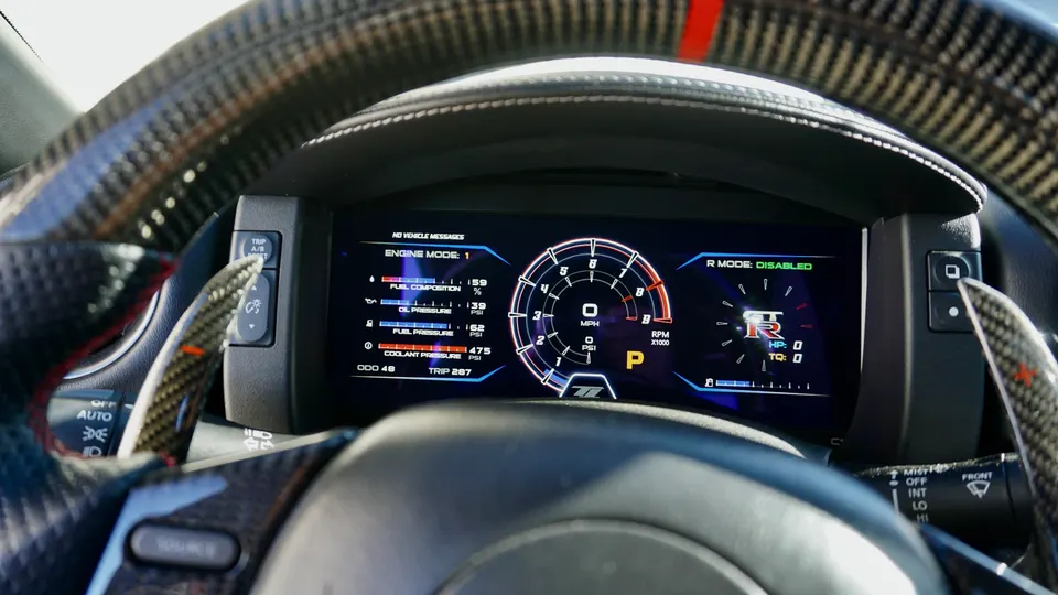 A close-up of a Nissan GT-R's digital dashboard displaying engine data and a carbon fiber steering wheel with paddle shifters.