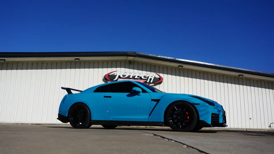 A bright blue Nissan GT-R with black wheels parked in front of the Jotech Motorsports building on a sunny day.