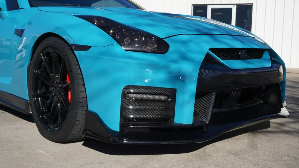 A blue Nissan GT-R with carbon fiber front bumper and splitter, black aftermarket wheels, and red brake calipers.