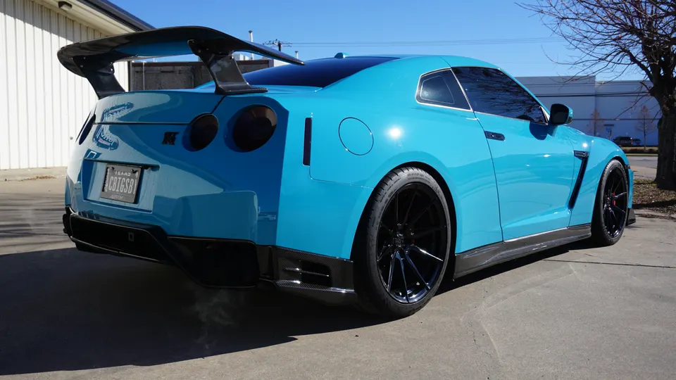 A blue Nissan GT-R with a large carbon fiber wing, aftermarket wheels, and carbon fiber aero parts parked outdoors.