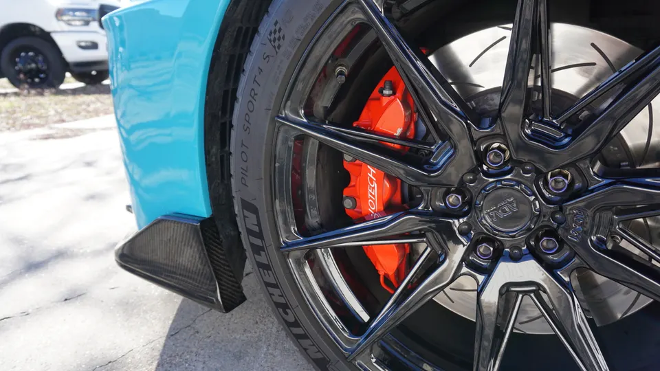 Close-up of a turquoise car's black ADV.1 wheel, Michelin Pilot Sport 4 S tire, and red Brembo brake caliper with a carbon fiber aero piece.