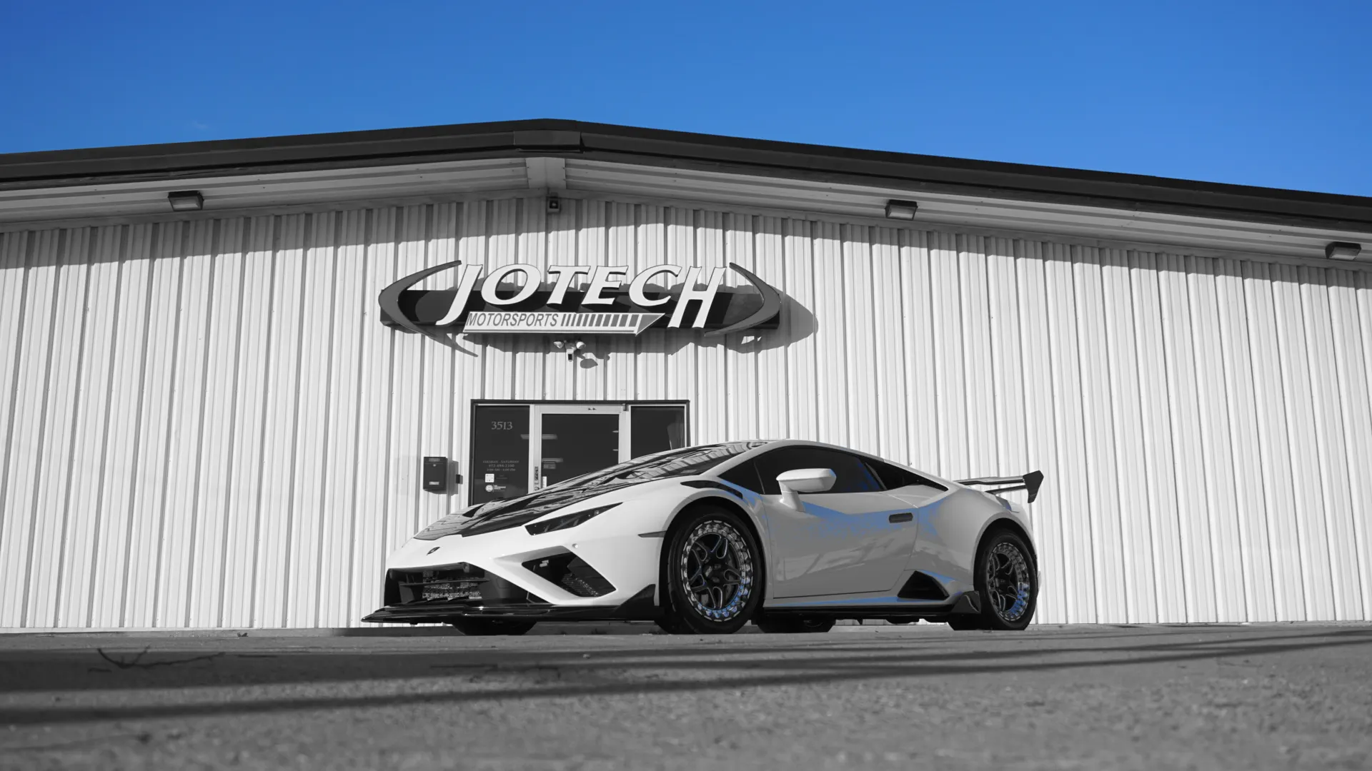 A white Lamborghini Huracan with aftermarket wheels and an aero kit parked in front of the Jotech Motorsports shop.