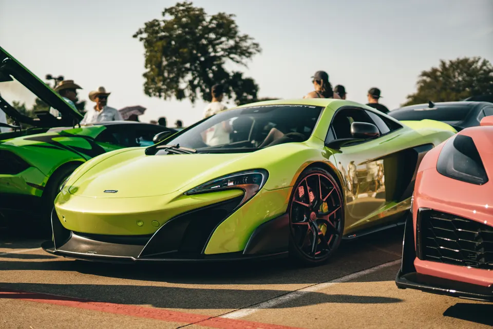 A vibrant lime green McLaren supercar parked at an outdoor car show, with other luxury vehicles and people in the background.