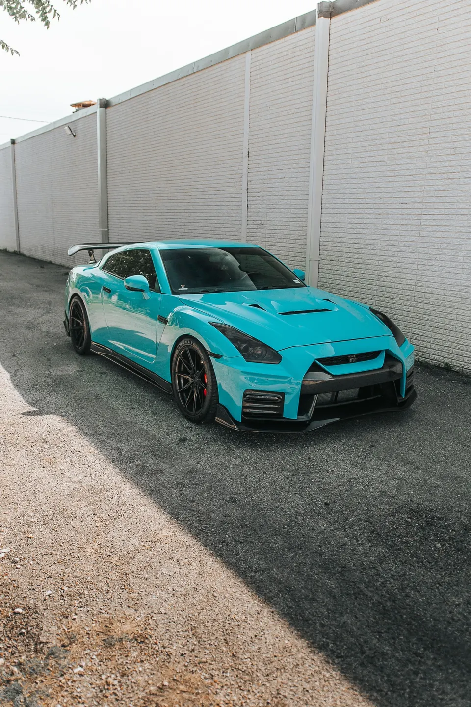A turquoise Nissan GT-R with aftermarket wheels and carbon fiber aero parked in an alley next to a white brick wall.