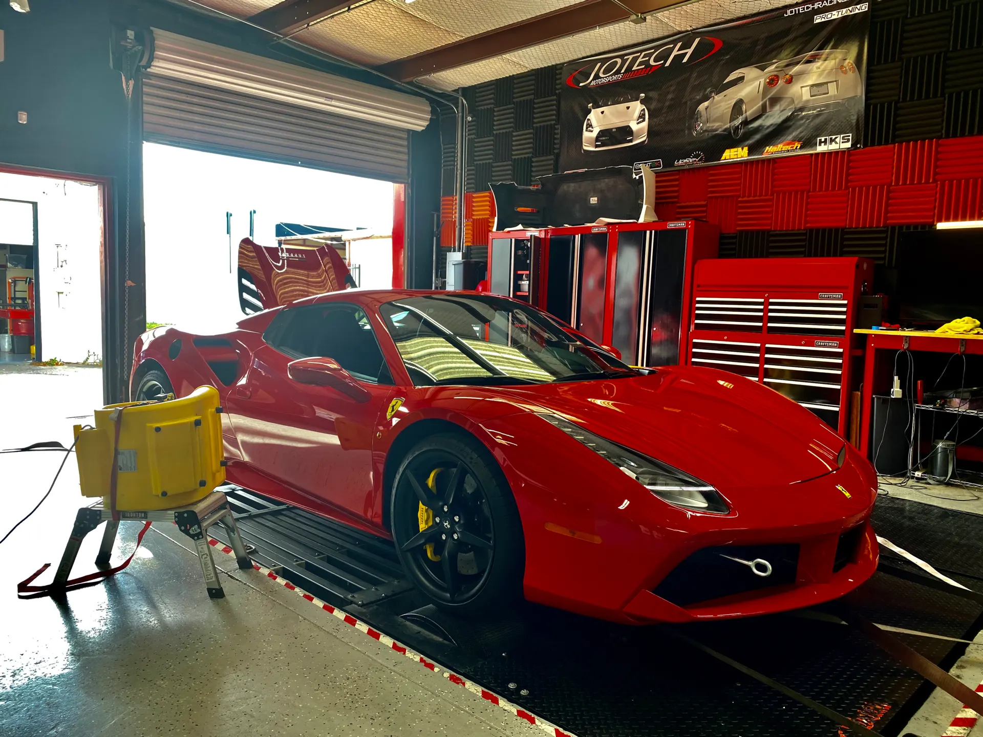 A red Ferrari 488 supercar strapped to a dynamometer for performance tuning at a Jotech Motorsports shop.
