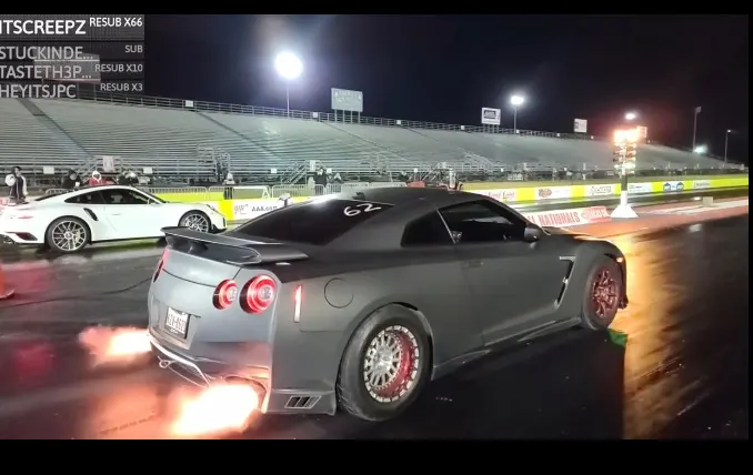 A Nissan GT-R with exhaust flames launching at a drag strip next to a Porsche 911 at night.