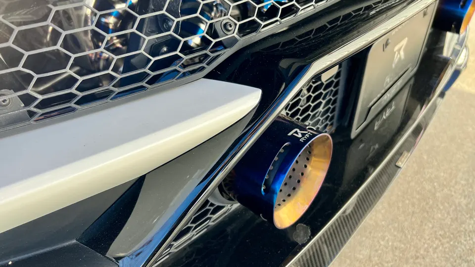 A close-up of a blue and gold titanium exhaust tip on a performance car, integrated into the bumper.