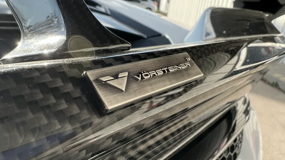 Close-up of a carbon fiber Vorsteiner rear wing on a performance car, showcasing the intricate weave and badge.