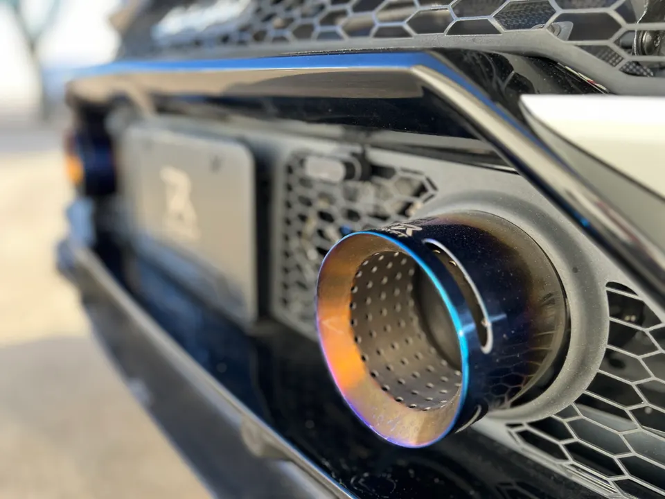 Close-up of a performance exhaust tip with a burnt titanium finish on a car's rear bumper, featuring a honeycomb grille.