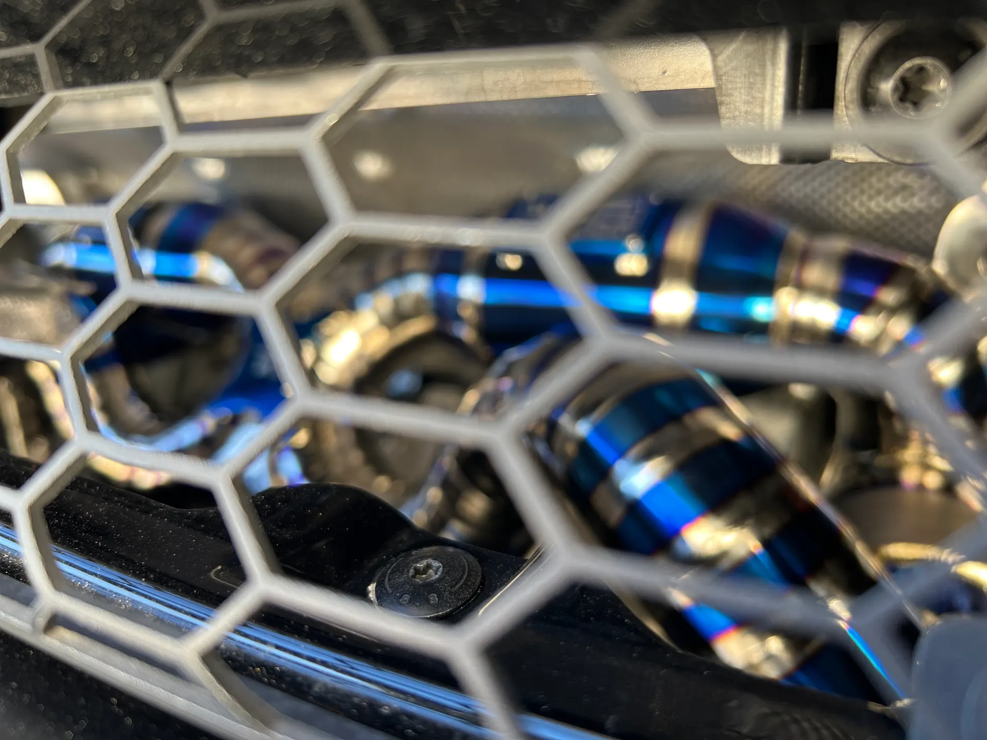 A close-up detail shot of a heat-treated titanium exhaust pipe with blue and gold hues, visible through a honeycomb grille.