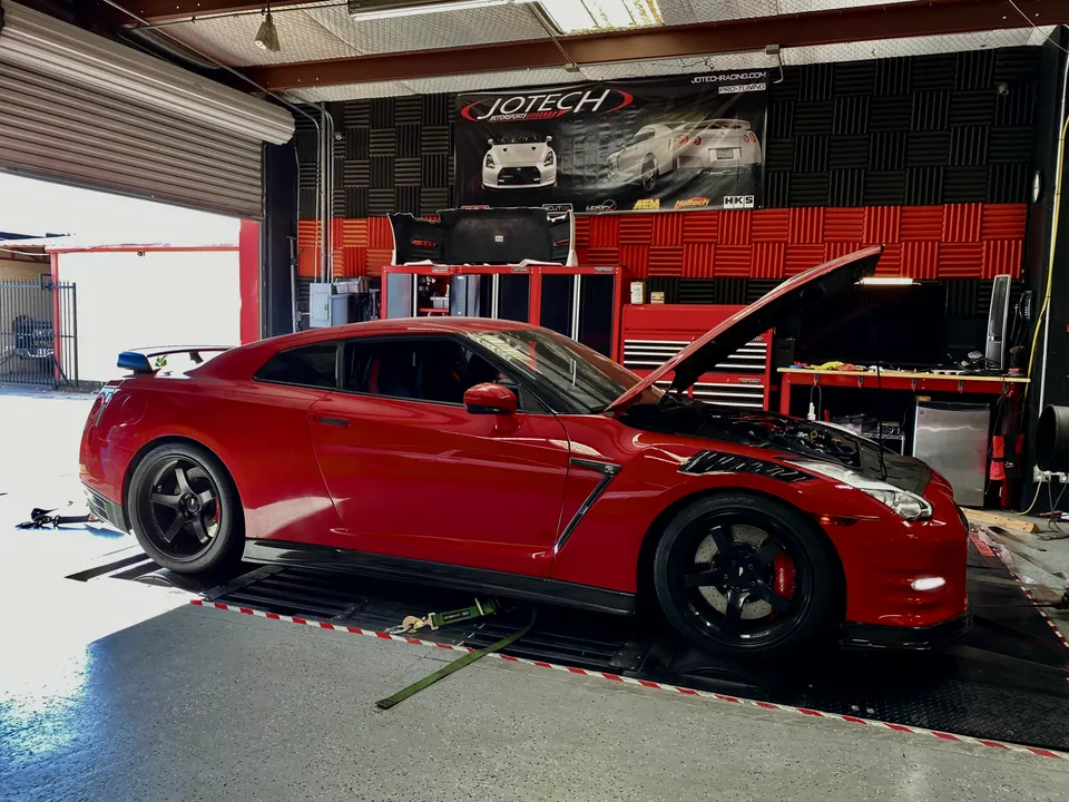 A red Nissan GT-R R35 with its hood open, strapped to a dynamometer in a Jotech Motorsports tuning shop.