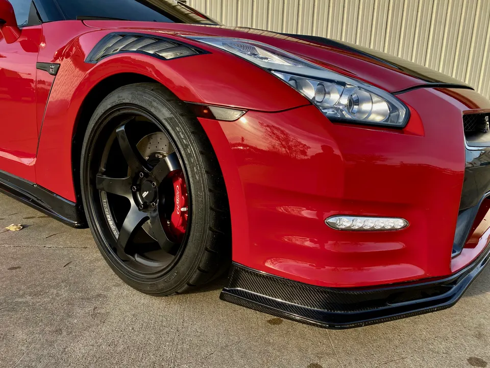 Close-up of a red Nissan GT-R's front wheel, showing black aftermarket wheels, red Brembo calipers, and a carbon fiber front splitter.