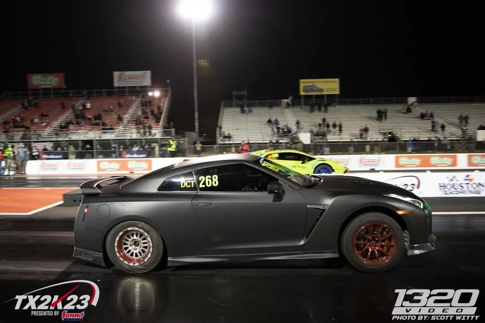 A dark grey Nissan GT-R with aftermarket wheels on a drag strip at night, with a yellow Lamborghini in the background.